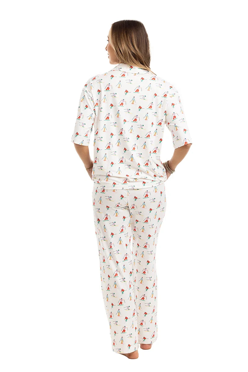 A woman stands barefoot facing away, wearing the Katydid Apres Ski Matching Pajama Pants Set with a colorful holiday print, featuring a short-sleeve top and loose-fitting long pants against a plain white background.