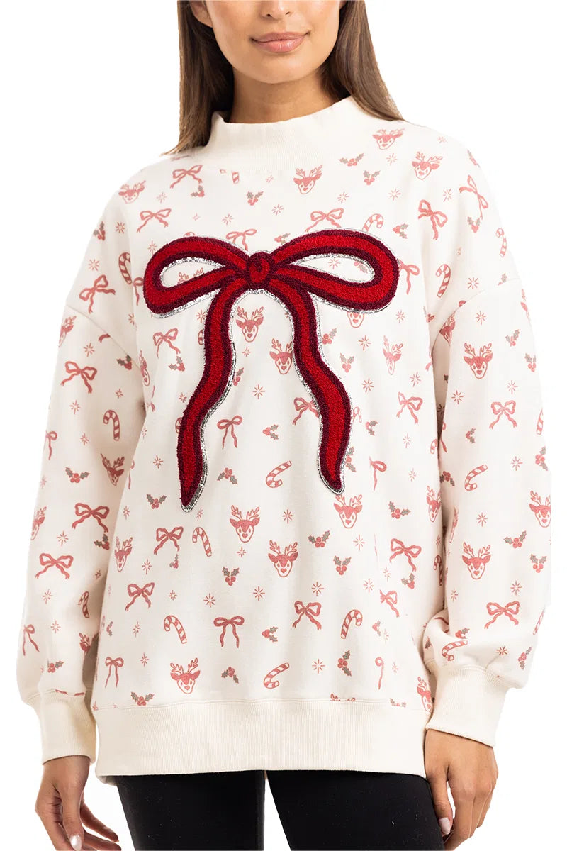 A woman wears the Katydid Bow Patch Reindeer Mockneck Sweatshirt, featuring festive reindeer faces, candy canes, holly, and a large textured red bow patch on the front.