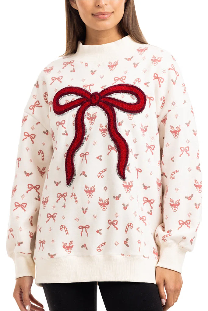 A woman wears the Katydid Bow Patch Reindeer Mockneck Sweatshirt, featuring festive reindeer faces, candy canes, holly, and a large textured red bow patch on the front.