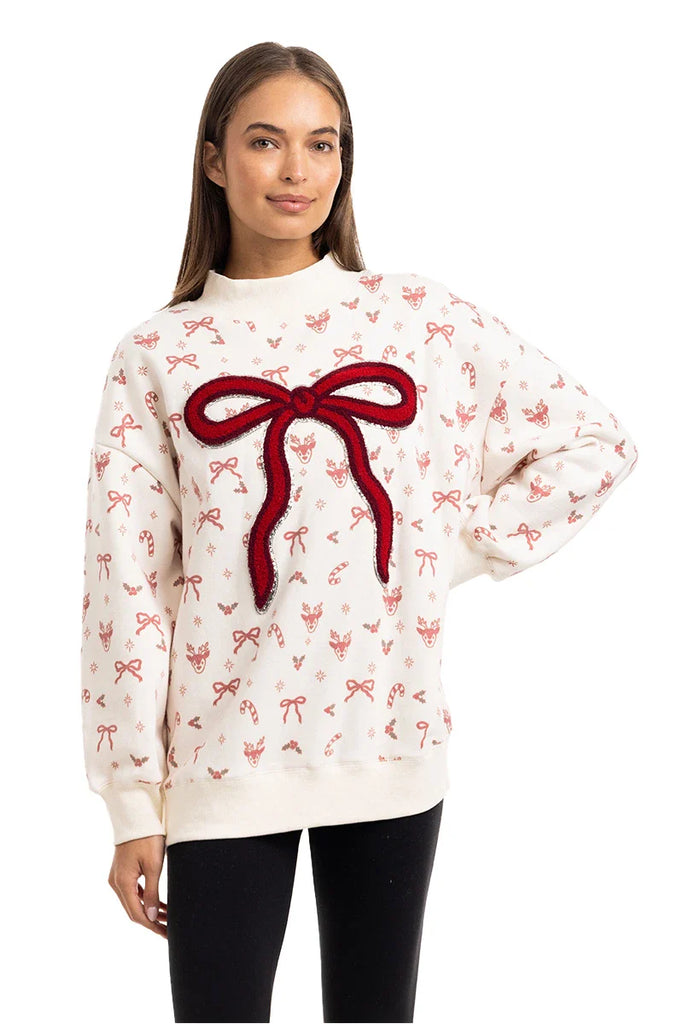 A woman wears the Katydid Bow Patch Reindeer Mockneck Sweatshirt, featuring a large red bow patch and scattered small bows with floral designs, stylishly paired with black leggings.