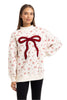 A woman wears the Katydid Bow Patch Reindeer Mockneck Sweatshirt, featuring a large red bow patch and scattered small bows with floral designs, stylishly paired with black leggings.