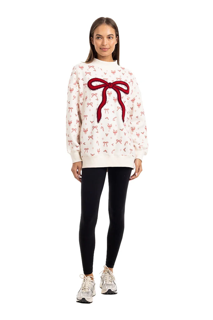 A woman wears the Katydid Bow Patch Reindeer Mockneck Sweatshirt, featuring a large red bow patch and smaller bow patterns, paired with black leggings and white sneakers. She smiles against a white background.