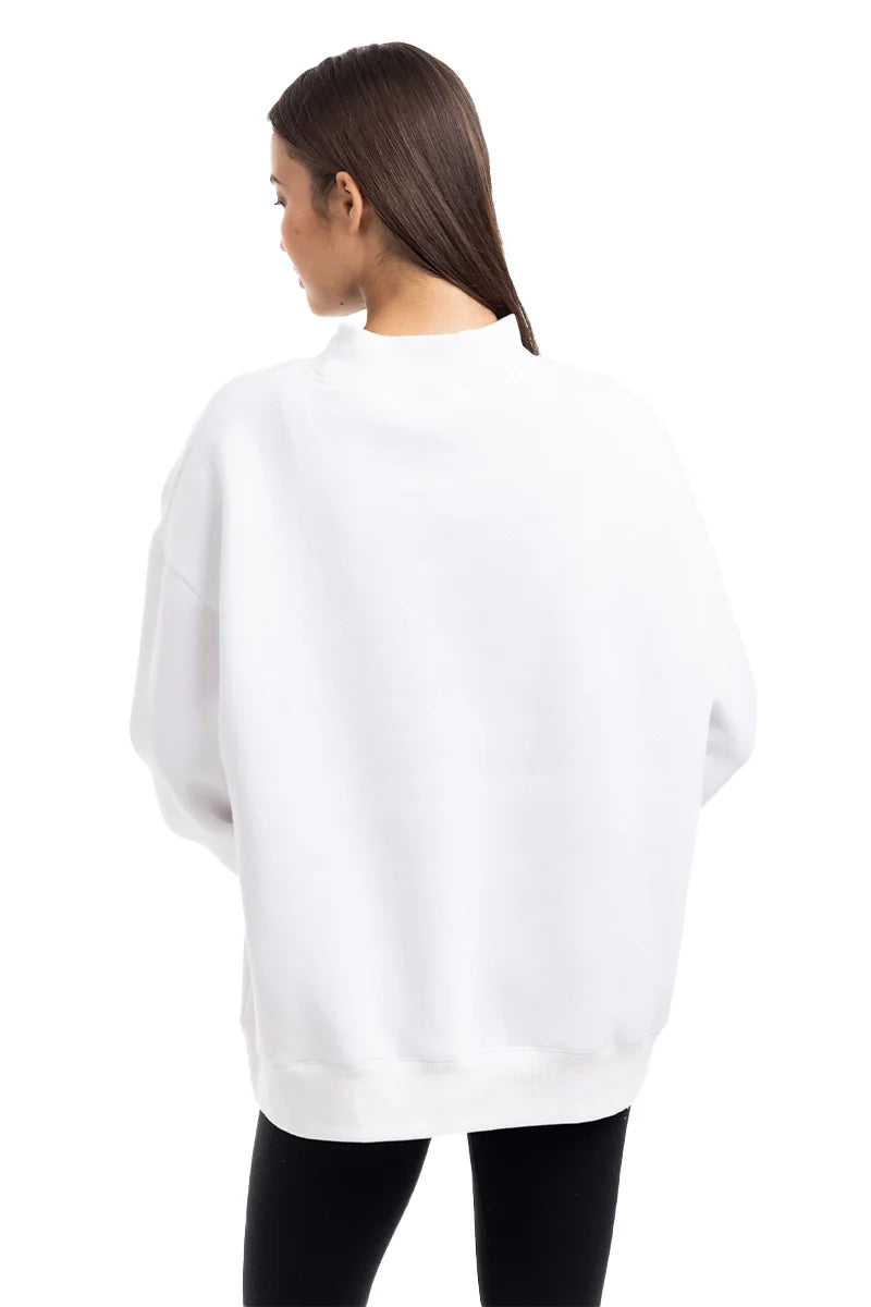 A woman with long brown hair, wearing the Katydid Cloud White Essential Mockneck Sweatshirt and black pants, stands facing away from the camera against a plain white background.