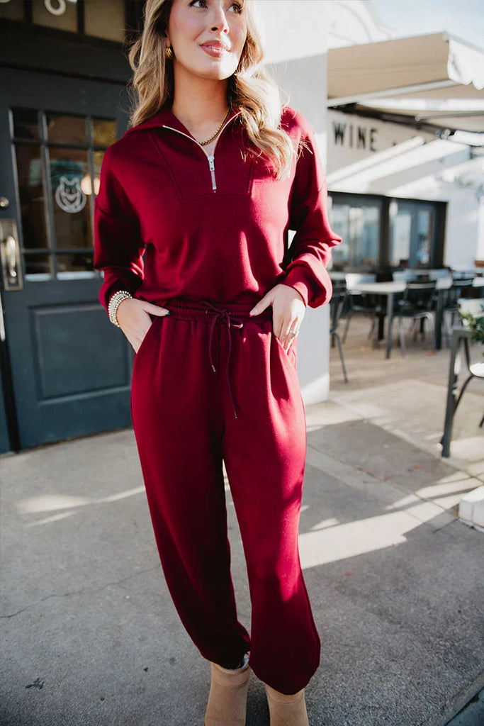 A woman smiles with one hand in her pocket, wearing the Katydid Wine Pants Two-Piece Set—a matching maroon long-sleeve zip-up top and drawstring pants—against an inviting outdoor café backdrop.