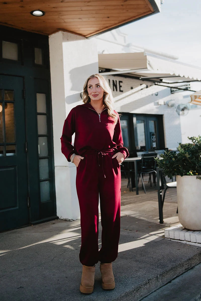 Wearing the Katydid Wine Pants Two-Piece Set and tan platform boots, a woman stands outside a cafe, smiling with her hands in her pockets, surrounded by tables, chairs, and potted plants on the sidewalk.