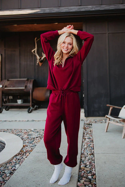 A smiling person in the Katydid Wine Pants Two-Piece Set poses outdoors on a stone path, arms raised, beside a grill and patio chair.