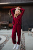 A smiling person in the Katydid Wine Pants Two-Piece Set poses outdoors on a stone path, arms raised, beside a grill and patio chair.