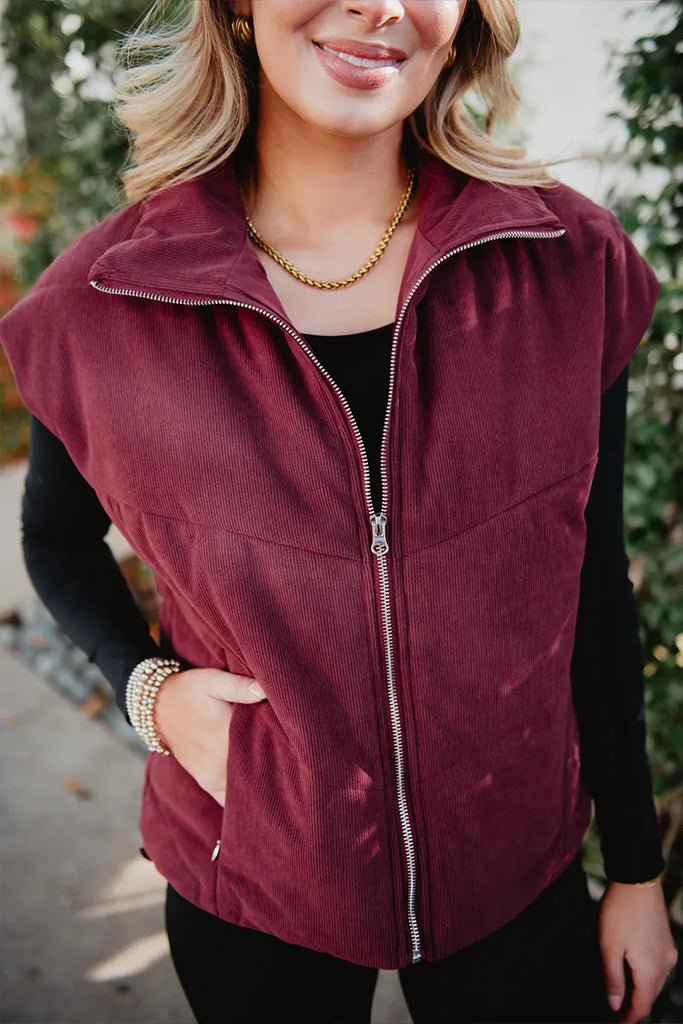 A woman models the Katydid Wine Fall Corduroy Puffer Vest over a black long-sleeve top with gold jewelry, standing outdoors among greenery and highlighting chic fall style.