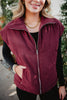 A woman models the Katydid Wine Fall Corduroy Puffer Vest over a black long-sleeve top with gold jewelry, standing outdoors among greenery and highlighting chic fall style.