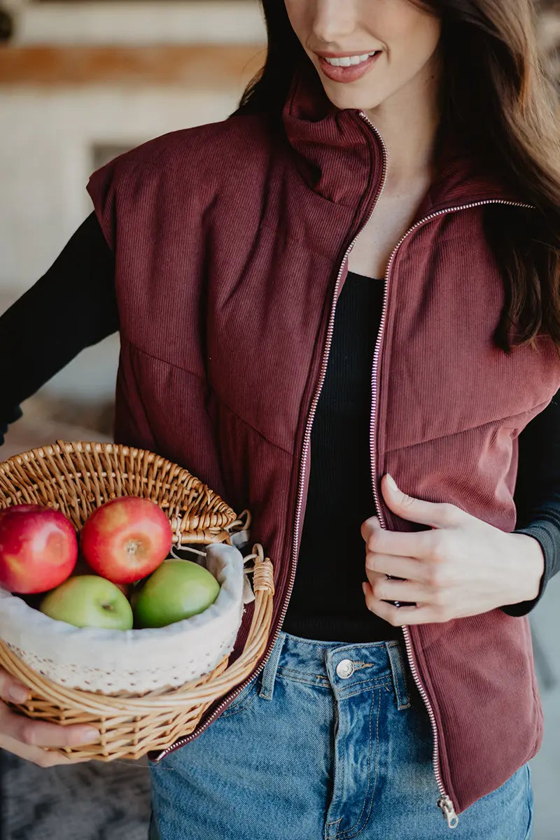 A woman wears the Katydid Wine Fall Corduroy Puffer Vest with jeans, holding a wicker basket of apples. She smiles indoors, showcasing cozy fall style with only her upper body visible.