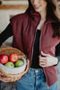 A woman wears the Katydid Wine Fall Corduroy Puffer Vest with jeans, holding a wicker basket of apples. She smiles indoors, showcasing cozy fall style with only her upper body visible.