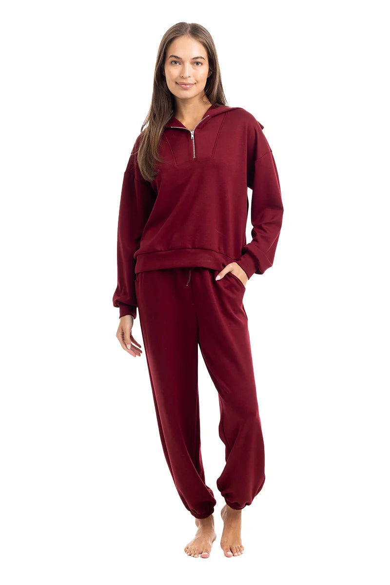 A woman smiles barefoot in the Katydid Wine Pants Two-Piece Set, featuring a cozy zip-up pullover top and jogger pants, posed against a white background.
