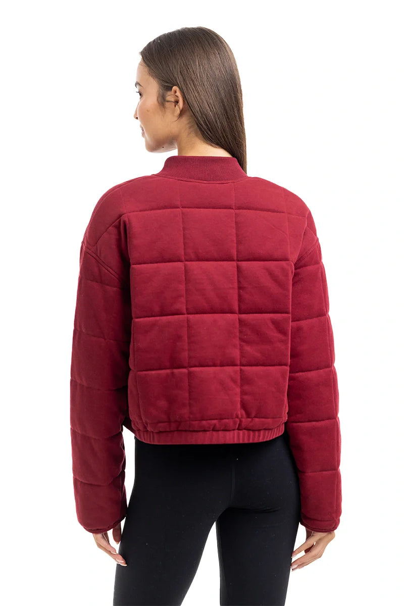 A woman with long brown hair, seen from the back, models the Katydid Womens Wine Cropped Quilted Bomber Jacket paired with black leggings against a white background.