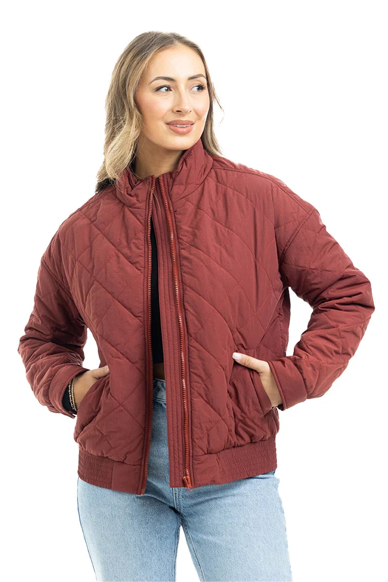 A woman with light brown hair smiles as she stands, hands in pockets, wearing the Katydid Wine Diamond Quilted Jacket and light blue jeans against a white background.