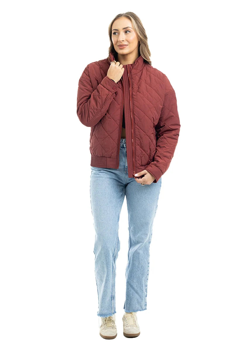 A woman stands against a white background wearing the Katydid Wine Diamond Quilted Jacket with light blue jeans and beige sneakers. She has long hair, smiles slightly, and holds the jacket near her collar with one hand.