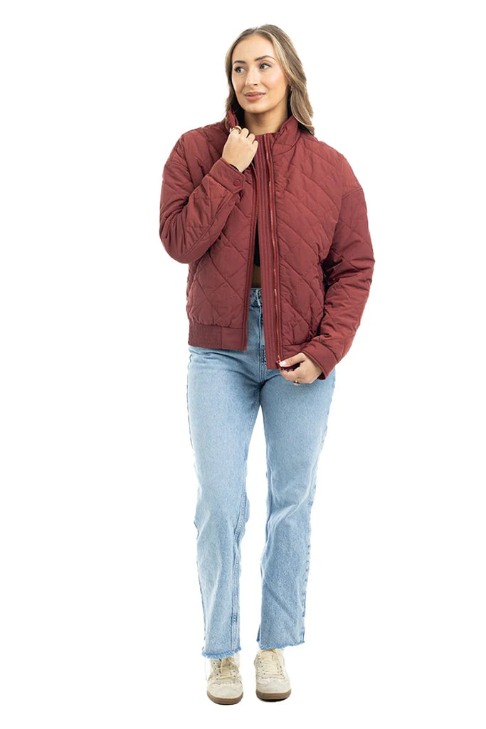 A woman stands against a white background wearing the Katydid Wine Diamond Quilted Jacket with light blue jeans and beige sneakers. She has long hair, smiles slightly, and holds the jacket near her collar with one hand.