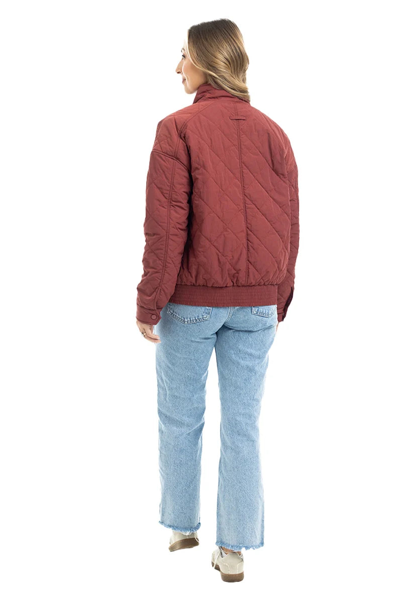 A person with long hair, wearing the Katydid Wine Diamond Quilted Jacket, light blue jeans, and beige shoes, stands facing away from the camera against a white background.