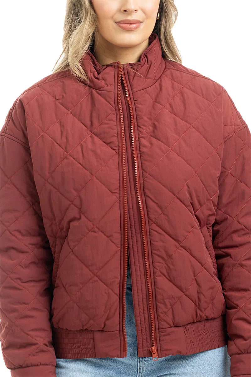 A person wears the Katydid Wine Diamond Quilted Jacket—a maroon zip-front womens jacket with a high collar—paired with light blue jeans, standing against a white background.