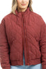 A person wears the Katydid Wine Diamond Quilted Jacket—a maroon zip-front womens jacket with a high collar—paired with light blue jeans, standing against a white background.