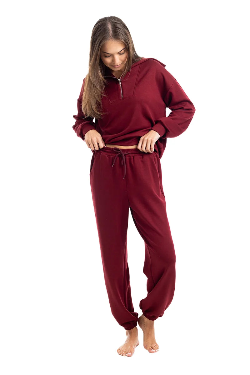 A woman models the Katydid Wine Pants Two-Piece Set, a cozy zip-up top paired with matching pants, standing barefoot and adjusting her outfit against a plain white background.