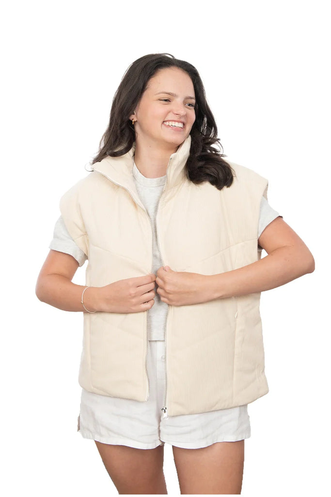 A woman smiles in Katydid’s Cloud White Corduroy Puffer Vest, styled over a gray t-shirt and white shorts. She stands against a plain white background, holding the vest with both hands.