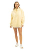 A woman smiles in a Katydid Yellow Vintage Wash Pullover Sweatshirt, styled with white shorts and sneakers against a plain white background.