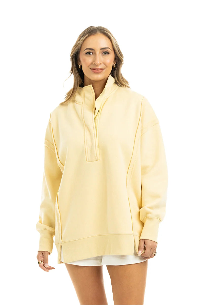 A woman with light brown hair stands smiling against a plain white background, wearing the Katydid Yellow Vintage Wash Pullover Sweatshirt and white shorts.