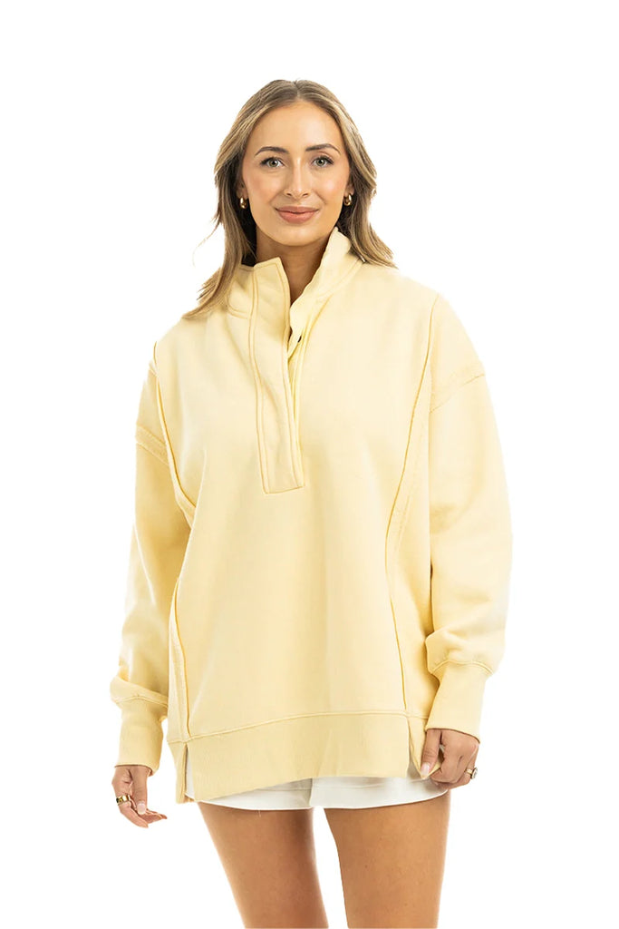 A woman with light brown hair stands smiling against a plain white background, wearing the Katydid Yellow Vintage Wash Pullover Sweatshirt and white shorts.