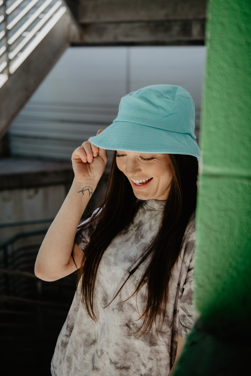 A smiling woman with long brown hair wears the Katydid Aqua Corded Bucket Hat, paired with a tie-dye shirt. She stands outdoors by a green wall with industrial accents, holding the brim to showcase this trendy boutique accessory.