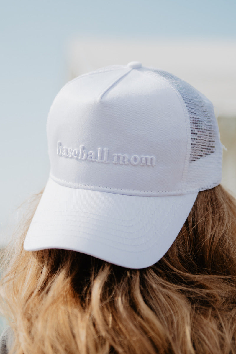White mesh baseball cap with "baseball mom" embroidered on the front, worn by a person with wavy brown hair, set against a clear sky.
