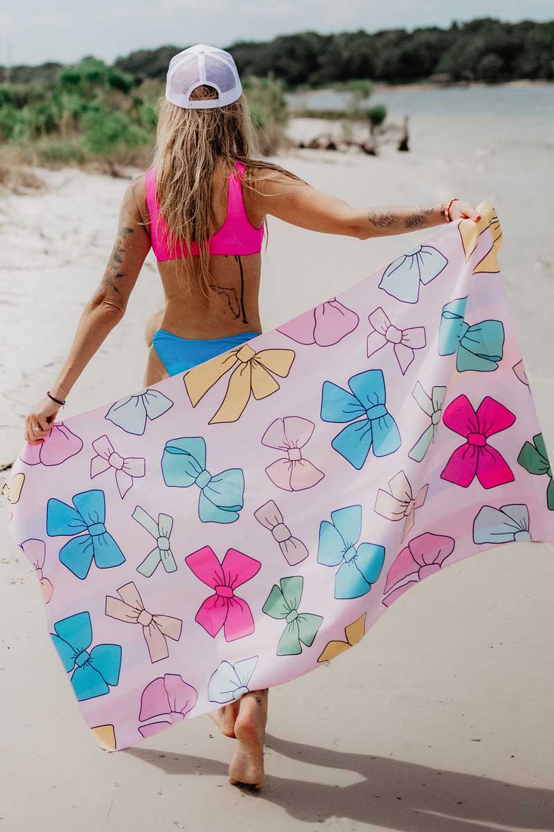 A woman in a pink bikini top and blue bottoms holds a colorful bow-patterned towel on a sandy beach, exuding a carefree, summery vibe.