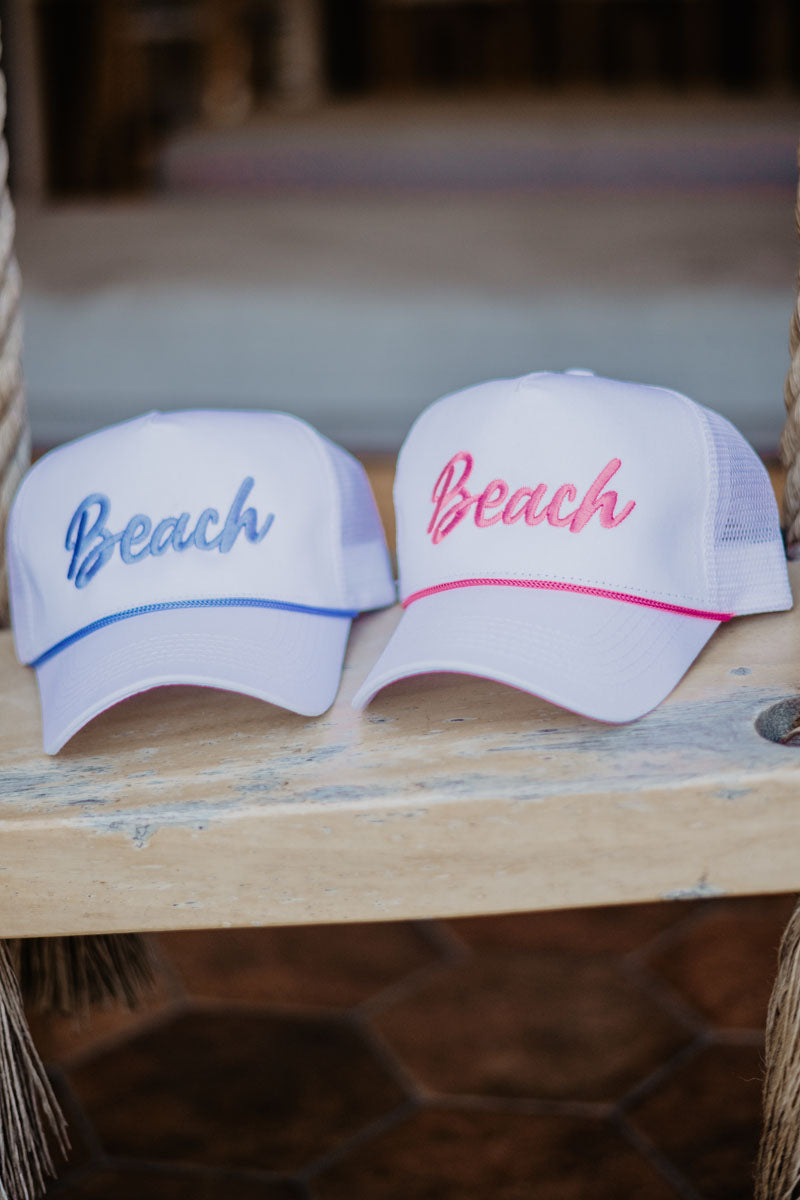 Two white caps with "Beach" embroidered in blue and pink sit on a wooden bench. The scene conveys a casual, relaxed, and summery vibe.