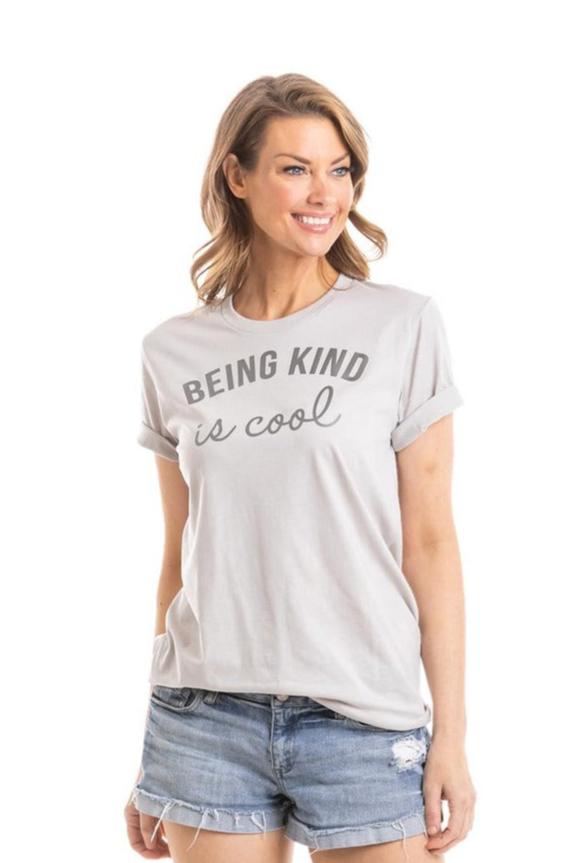 A woman smiles in the Katydid Being Kind is Cool Women's T-Shirt, a light gray round neck tee with rolled sleeves and bold print, styled with denim shorts against a plain white background for a modern casual look.