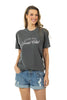 A woman stands smiling with one hand in her pocket against a white background, wearing Katydid’s Game Day Social Club Graphic Tee in dark gray paired with distressed denim shorts.
