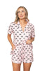 A woman with blonde hair models the Katydid Black  Pink Mini Coquette Bows Loungewear Set—featuring a button-up shirt and drawstring shorts with playful mini bows. She smiles slightly, hand in pocket, against a white background.