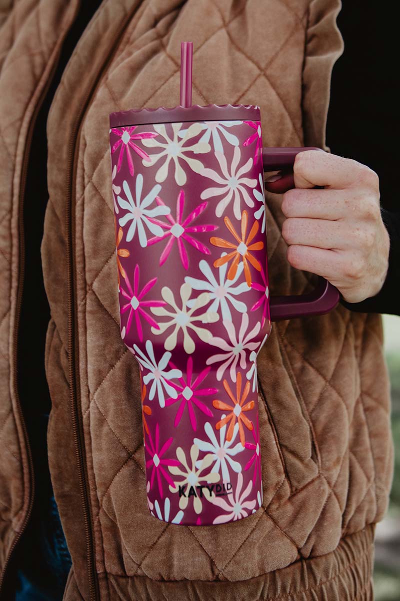 A person in a brown quilted jacket holds a large floral-patterned pink tumbler with a handle and straw. The design is vibrant and cheerful.
