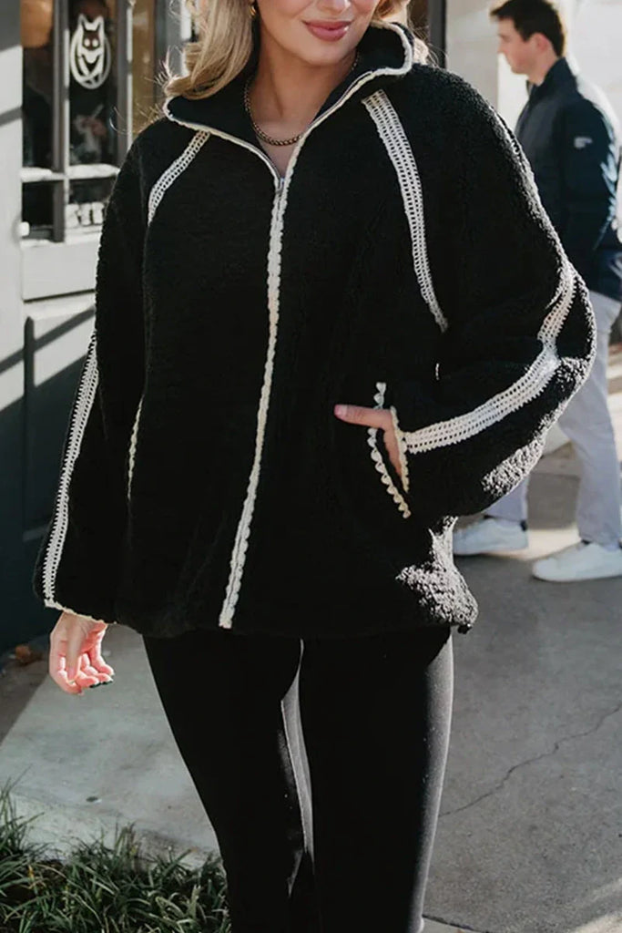 A woman wears the Katydid Black Sherpa Lace Trim Jacket with black leggings, standing outdoors with one hand in her pocket. Another person is visible in the background.