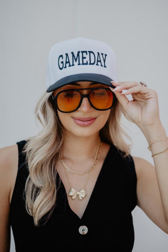 Young woman wearing a "GAMEDAY" cap and large sunglasses smiles slightly. Her wavy hair frames her face, and she wears layered necklaces with a bow charm.