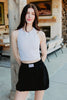 A woman with long dark hair stands outside near a wooden post, wearing a Katydid Black Bubble Skirt and a white sleeveless top, with outdoor furniture and stacked firewood in the background.