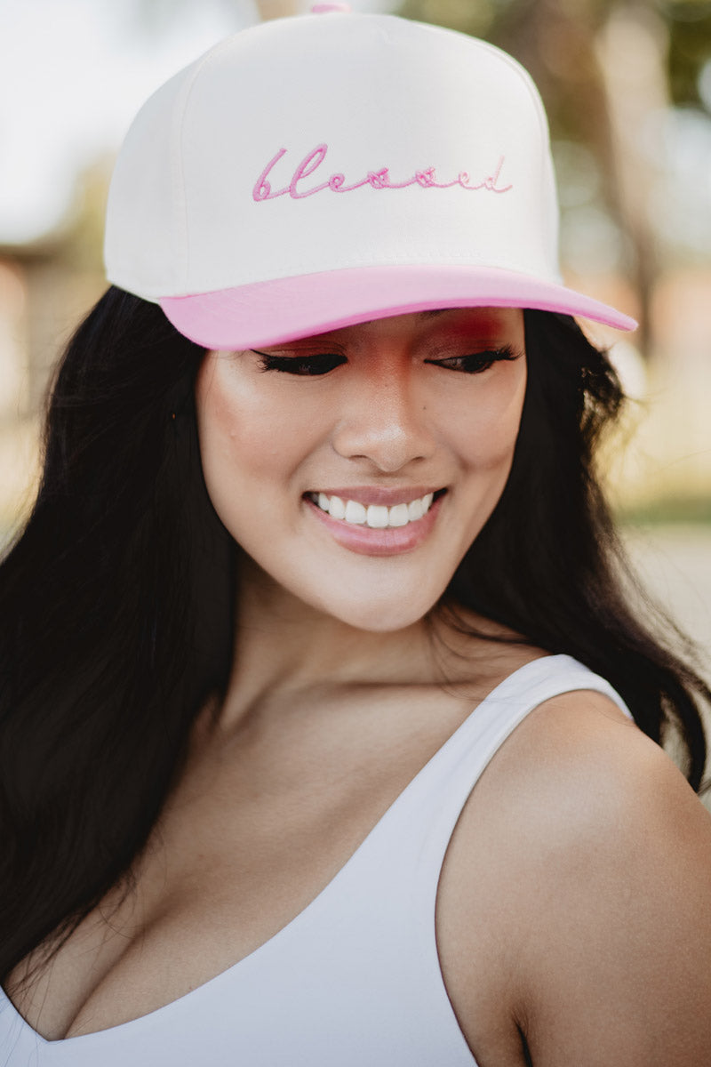 Smiling woman wearing a white and pink cap with "blessed" embroidered on it, stands outdoors. She exudes happiness and confidence.