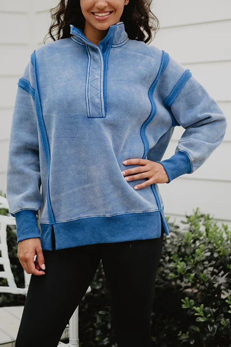 A woman in the Katydid Blue Vintage Wash Snap-Neck Pullover and black pants stands outdoors by green bushes and a white wall.
