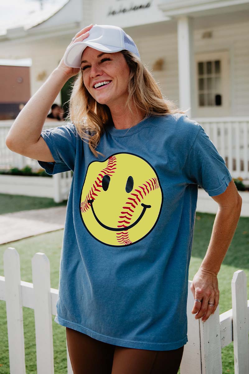 A smiling woman stands outdoors by a fence and house wearing the Katydid Softball Happy Face Graphic Tee—a blue, garment-dyed cotton T-shirt featuring a large yellow softball-inspired smiley face—and touches her white cap.