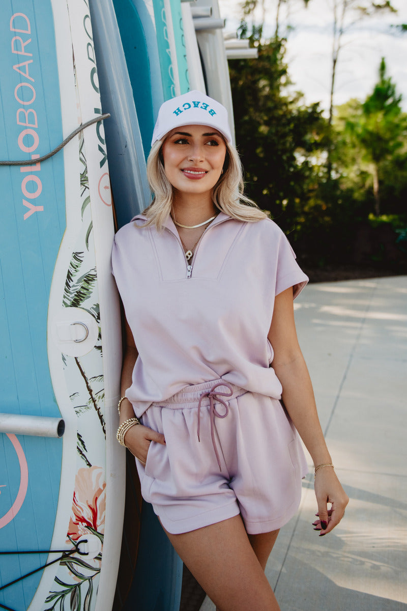 Smiling woman in a pastel outfit and "Beach" cap leans against paddleboards labeled "YOLOBOARD," set in a sunny outdoor environment.