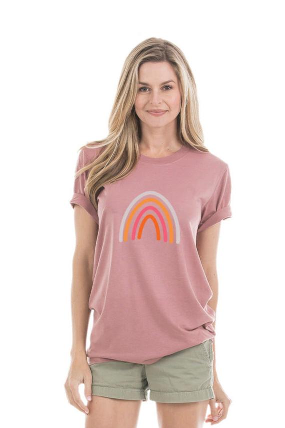 A woman with long blonde hair smiles at the camera, wearing the Katydid Boho Rainbow Graphic Tee for women in mauve with a pink and orange Nordic rainbow design, paired with light green shorts. She stands against a plain white background.
