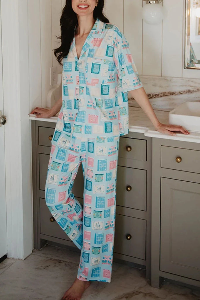 A person stands barefoot in a bathroom, wearing Katydid’s Book Club Matching Pajama Sets featuring a colorful graphic pattern. They lean against a marble countertop, with gray cabinets and white paneled walls behind them.