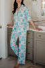 A person stands barefoot in a bathroom, wearing Katydid’s Book Club Matching Pajama Sets featuring a colorful graphic pattern. They lean against a marble countertop, with gray cabinets and white paneled walls behind them.