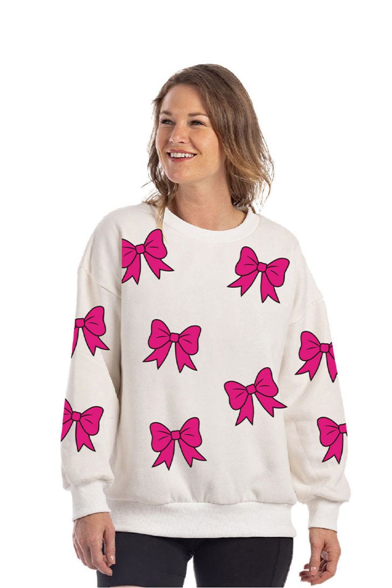 A woman smiles in the Katydid Sequined Coquette BOW Patches Sweatshirt, featuring large pink sequin bows. The oversized cotton-polyester top flatters her light brown hair as she poses against a plain background.