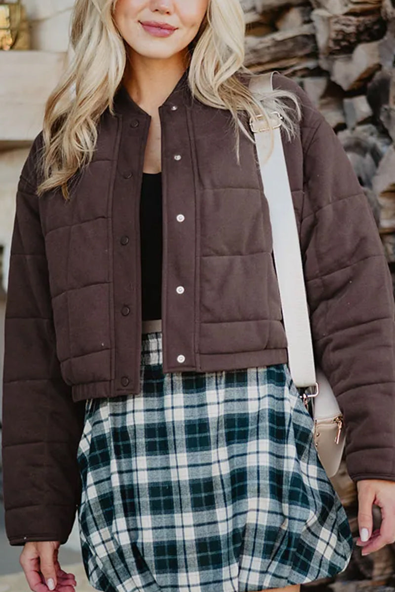 A woman with loose, wavy blonde hair stands outdoors carrying a light-colored shoulder bag, wearing the Katydid Chocolate Brown Cropped Quilted Bomber Jacket over a black top and blue and white plaid skirt.