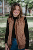 A young woman smiles outdoors in a black long-sleeve top, blue jeans, and the Katydid Brown Garment Washed Quilted Ruffle Sleeve Vest, embracing a cozy fall vibe with trees and a building blurred in the background.