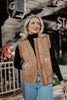 A woman with long blonde hair smiles outside on a sunny day, wearing a black turtleneck, blue jeans, and the Katydid Brown Garment Washed Quilted Ruffle Sleeve Vest, capturing a cozy fall vibe.